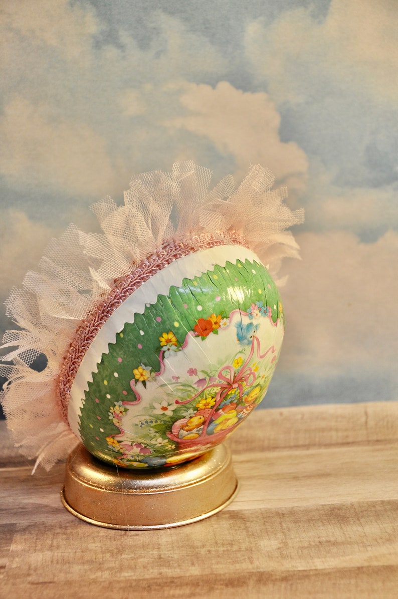 May include: A green and white Easter egg with a pink tulle ruffle and a floral design. The egg is sitting on a gold stand.