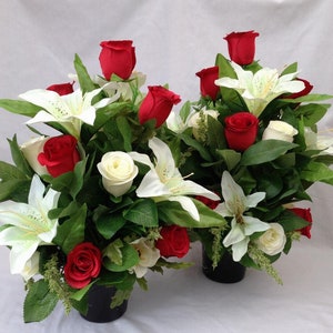 May include: Two artificial flower arrangements in black pots, featuring white lilies and red roses.