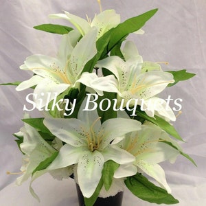 May include: A white silk lily bouquet in a black pot with a white base. The bouquet is arranged in a vase with green leaves. The text "Silky Bouquets" is visible in the image.