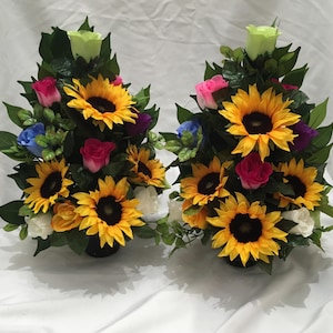 May include: Two artificial flower arrangements in black pots. Each arrangement features a large yellow sunflower, surrounded by smaller sunflowers, pink, white, blue, and purple roses, and green leaves.