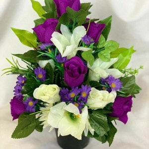 May include: An artificial flower arrangement in a black pot, featuring purple and white roses, lilies, and smaller purple flowers.