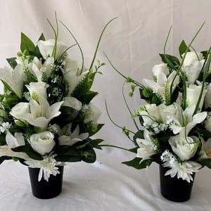 May include: Two floral arrangements with white roses, lilies, and other white flowers with green leaves. The flowers are in black pots. The arrangements are set against a white background.