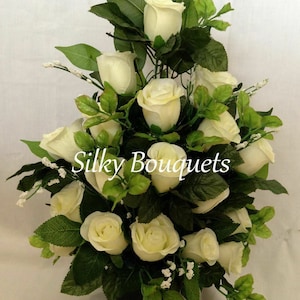 May include: A white silk flower arrangement with a variety of white roses and greenery in a black vase. The arrangement is labelled "Silky Bouquets".