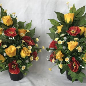 May include: Two artificial flower arrangements in black pots. Each arrangement features yellow roses, red poppies, and white daisies.