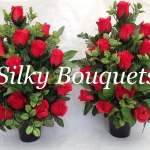 May include: Two artificial red rose bouquets in black pots. The bouquets are arranged in a cascading style with green leaves. The text "Silky Bouquets" is printed in white on a black background.