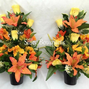 May include: Two artificial flower arrangements in black pots. Each arrangement features orange lilies, yellow roses, and orange and yellow flowers. The arrangements are on a white background. Silky Bouquets is written in the center of the image.