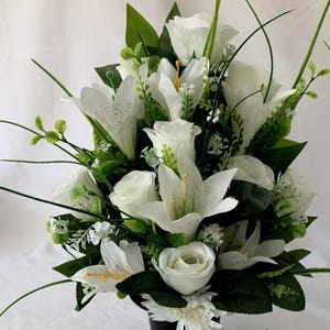 May include: A white artificial flower arrangement in a black vase. The arrangement features white roses and lilies with green foliage.