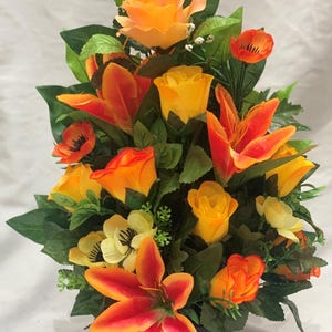 May include: A silk flower arrangement in a black pot. The arrangement features orange roses, lilies, and poppies.