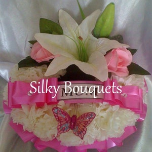 May include: A white and pink flower arrangement featuring a white lily, pink roses, and white carnations. The arrangement is heart-shaped and has a pink ribbon with the words "Silky Bouquets" and "Mum" on it. A sparkly butterfly is in the centre of the arrangement.