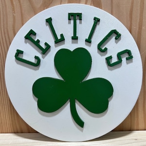 May include: A white circular wooden sign with a green shamrock and the word "CELTICS" in black letters.