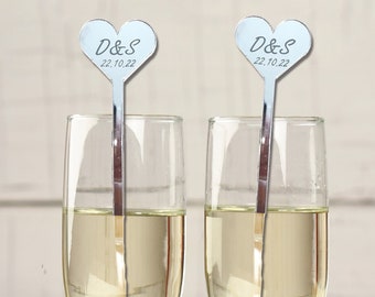 May include: Two champagne flutes with silver heart-shaped drink stirrers. The stirrers have the text "D&S 22.10.22" on them. The flutes are surrounded by white roses.