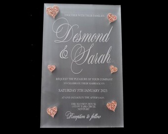 May include: A clear acrylic wedding invitation with the names Desmond & Sarah printed in white script. The invitation features a pink glitter heart design and the text "Together with their families". The date of the wedding is Saturday 5th January 2023, at one o'clock in the afternoon. The location is The Mansion House, 1 Stow Park Circle, Newport. The invitation concludes with "Reception to follow".