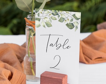 May include: A white card with the text "Table 2" printed on it. The card is propped up on a small wooden block. A small vase with a white flower and green leaves is in front of the card.