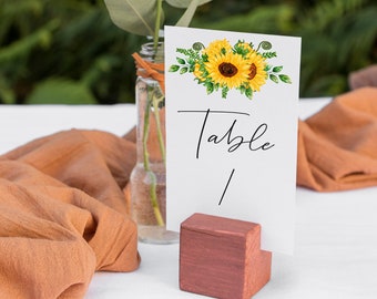 May include: A white card with a sunflower design and the text "Table 1" is displayed on a wooden stand. The card is propped up against a clear glass vase with a white flower and green leaves.