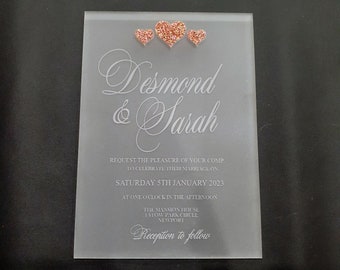 May include: A frosted acrylic wedding invitation with the names "Desmond & Sarah" in elegant script. Three rose gold glitter hearts adorn the top. The invitation includes the date, time, and location of the wedding.