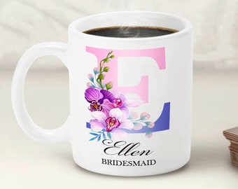May include: White ceramic mug with a pink and blue floral design and the letter "E" in pink. The mug also has the text "Ellen Bridesmaid" in black.