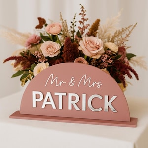 Puede incluir: Un letrero de madera color rosa polvoriento con las palabras "Mr & Mrs PATRICK" en letras plateadas. El letrero está frente a un arreglo floral de rosas y otras flores en tonos rosa, crema y burdeos.