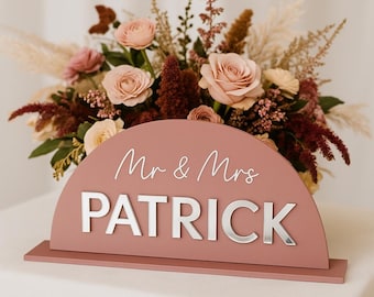 May include: A dusty rose-colored wooden sign with the words "Mr & Mrs PATRICK" in silver lettering. The sign is in front of a floral arrangement of roses and other flowers in shades of pink, cream, and burgundy.
