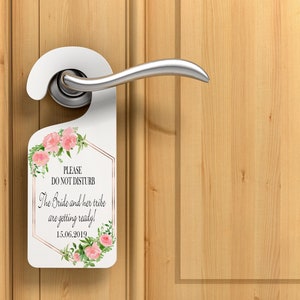 Puede incluir: Un colgador de puerta blanco con un dise&ntilde;o floral y el texto "PLEASE DO NOT DISTURB". Tambi&eacute;n se muestra el texto "The Bride and her tribe are getting ready! 15.06.2019". El colgador est&aacute; en una puerta de madera.