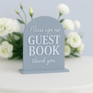 Puede incluir: Un letrero gris en forma de arco para el libro de visitas con el texto "Please sign our GUEST BOOK thank you" en blanco. El letrero está sobre una superficie blanca, con flores blancas y vegetación en el fondo.