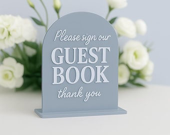 May include: A gray, arched guest book sign with the text "Please sign our GUEST BOOK thank you" in white. The sign is on a white surface, with white flowers and greenery in the background.