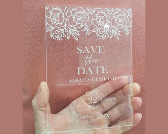 May include: A clear acrylic save the date card with white floral designs at the top. The text reads "Save the Date" with the names "Sarah & Desmond" below. The date is "Oct 22, 2022" with the time "1:30 PM" and the location "Caldicot Castle".