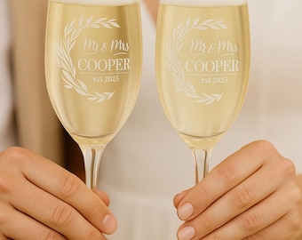 May include: Two champagne flutes filled with bubbly liquid, held by hands. Each glass is etched with "Mr & Mrs Cooper est. 2025" within a decorative wreath. The glasses are clear, and the liquid is a pale yellow.