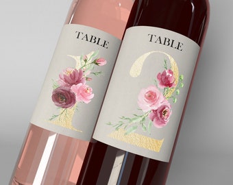 May include: Two wine bottles with labels featuring gold numbers and the word "TABLE". The numbers are adorned with watercolor floral arrangements in shades of pink and burgundy. One bottle contains a pink liquid, the other a dark red liquid.