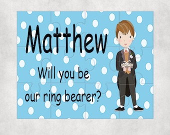 May include: Blue puzzle piece with white polka dots. The text "Matthew Will you be our ring bearer?" is on the puzzle piece. A cartoon boy in a tuxedo is holding a ring box.