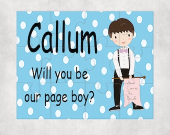 Op de afbeelding: Een blauwe puzzelstuk met witte stippen en de tekst "Callum Will you be our page boy?" en een cartoonillustratie van een jongen in pak die een bord vasthoudt met de tekst "Here Comes The Bride".