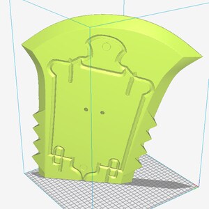 May include: A light green, 3D-printed object, possibly a mold or a component. It features a stylized, shield-like shape with a curved top and a series of jagged edges along one side. The design includes recessed details and two small circular holes.