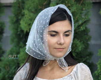 May include: A white sheer headscarf with small white polka dots. The scarf is tied in a bow at the back of the neck.
