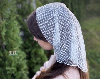 May include: A white lace head covering with a delicate pattern. The head covering is worn by a person with long brown hair.