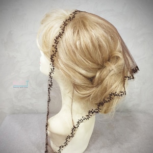 May include: A brown sheer veil with a delicate floral lace trim. The veil is worn on a mannequin with blonde hair styled in a bun.