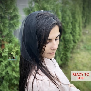 May include: A black sheer head covering with a small white embellishment. The head covering is worn by a person with long dark hair and a pink shirt.