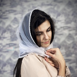 May include: A woman with long dark hair wears a white sheer veil with a delicate lace trim. She is wearing a beige long-sleeved shirt and has her hands clasped in front of her.