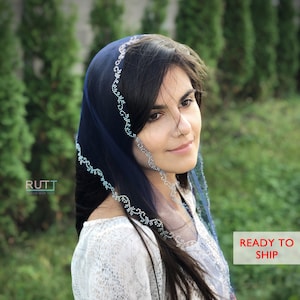 May include: A woman wearing a blue sheer headscarf with a beaded trim. The headscarf is draped over her head and shoulders. She is wearing a white blouse with a snake skin pattern. The background is a blurred image of green foliage.