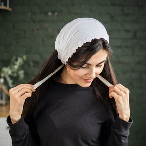 May include: A white headscarf with a beaded trim. The scarf is tied around the head and the ends are hanging down the sides.