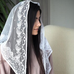 May include: A white lace veil with a paisley design. The veil is worn over a woman's head and shoulders.