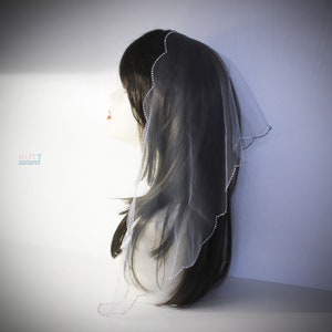 May include: A white tulle veil with a scalloped edge and a row of rhinestones. The veil is worn on a mannequin with long black hair.