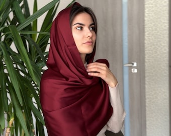 May include: A burgundy satin headscarf draped over the head and shoulders. The head covering is a deep, rich red color. The person is wearing a cream-colored sweater and sitting in a chair. A large green plant is in the background.