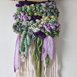 May include: A woven wall hanging with a driftwood top, showcasing a blend of textures and colours. The design incorporates purple, green, and white hues. Ribbons and yarn strands cascade from the bottom, creating a distinctive and artistic piece for home decor.