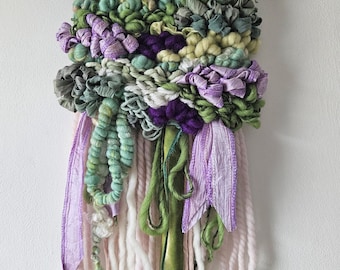 May include: A handmade textile wall hanging featuring a driftwood top and a woven design. The artwork incorporates various textures and colors, including shades of purple, green, and white. Ribbons and yarn strands hang from the bottom, adding to the piece's unique aesthetic.