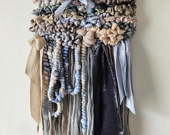 May include: A woven wall hanging with a natural wood branch at the top. The hanging features a variety of textures and colors, including beige, brown, blue, and gray. The hanging is made with yarn, fabric, and beads.