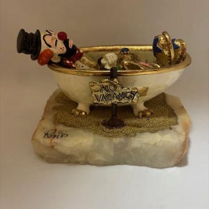 May include: A whimsical figurine of a clown relaxing in a bathtub. The tub is gold-rimmed and sits on a stone base. A sign reading "NO VACANCY" is in front of the tub. The clown wears a black top hat and red nose.