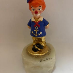 May include: A ceramic clown figurine in a sailor outfit. The clown has red hair, a white and blue sailor hat, and a blue shirt with a gold anchor. The figurine is standing on a white base with gold accents. Signed "Ron © 77".