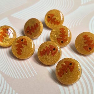 May include: A collection of eight circular buttons, each featuring a gold-toned, glittery resin base with a smooth, glossy finish. Embedded within each button is a distinct, orange-colored graphic of a fish skeleton, oriented in various directions.