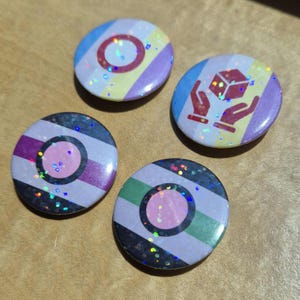 May include: Four circular pin-back buttons with a holographic glitter finish. Two buttons feature a central pink circle on a striped background of black, purple, and green. One button shows a red icon of hands holding a cube. One button has a red circle.