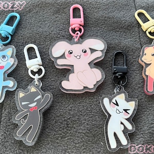 Op de afbeelding: Vijf kleurrijke sleutelhangers met cartoon dierenfiguren. De sleutelhangers zijn gemaakt van helder acryl en hebben een blauwe kat, een roze konijn, een witte kat, een zwarte kat en een perzikkleurige hond. Elke sleutelhanger heeft een metalen ring om sleutels aan te bevestigen.