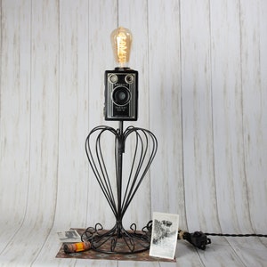 May include: A vintage camera lamp with a black and silver camera body, a clear glass bulb, and a black metal base with a heart-shaped design. The camera has the text "Brownie Target Six-16" on it.
